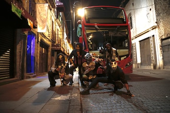 Cinco personas disfrazadas de monstruos posan frente a un autobús rojo de dos pisos en una calle empedrada durante la noche