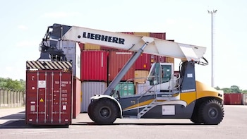 Una grúa apiladora de contenedores Liebherr de color gris y amarillo mueve un contenedor de carga rojo. Al fondo se ven más contenedores apilados