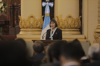 Mujer con chaqueta oscura y banda bicolor habla en un podio con micrófono. Detrás hay una bandera de Guatemala y decoración dorada