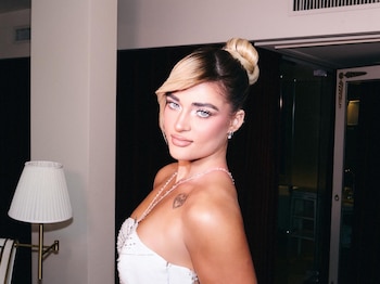 Marta Fort, de perfil, olhando para a câmera, usando vestido branco decotado, cabelos loiros e pretos presos e olhos azuis brilhantes.