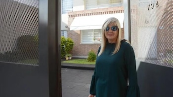Una mujer de cabello rubio con gafas de sol permanece junto a un portón abierto frente a un edificio de ladrillos