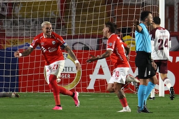 Un jugador de fútbol con camiseta roja número 21, Carlos Garcés, celebra un gol con un compañero en el campo de juego con una portería de fondo