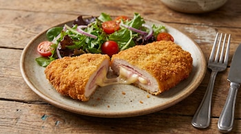 Milanesa de pollo empanada rellena de jamón y queso, cortada, con queso fundido. Acompaña ensalada de hojas verdes, tomates y cebolla morada, en plato sobre mesa de madera.