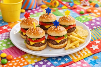 Plato blanco con cinco mini hamburguesas de queso, lechuga y tomate, acompañadas de patatas fritas y patatas chip, sobre un mantel de retazos colorido.
