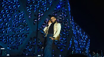 Hombre con barba, gafas y chaqueta bordada posa frente a un edificio moderno con fachada geométrica iluminada con luces azules en la oscuridad.
