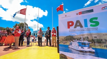 Personas en un estrado al aire libre con banderas peruanas y un cartel del Programa Nacional PAIS que exhibe una embarcación.