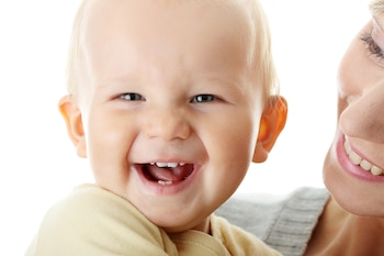 Primer plano de un bebé rubio sonriendo con la boca abierta y la parte inferior derecha del rostro de un adulto sonriendo, sobre un fondo blanco