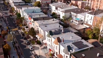 Vista aérea inclinada de una calle residencial flanqueada por casas adosadas de distintos estilos arquitectónicos, con coches estacionados y algunos tejados con paneles solares
