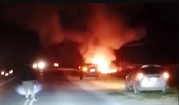 Vehículos aparcados de noche a un lado de la carretera mientras al fondo arde un fuego con humo denso