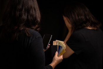 Vista posterior de una mujer con tarjetas, billetes y un teléfono móvil, mientras otra persona cubre su rostro al fondo.