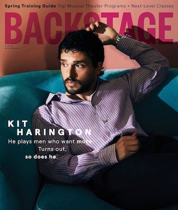 Kit Harington sentado en un sofá azul, con camisa de rayas moradas y oscuras, y barba. Mira a la izquierda con la mano en la cabeza. Fondo rosa con logo 'Backstage'