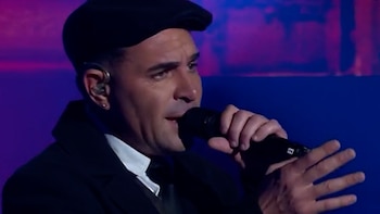 Primer plano de Performance M cantando en un micrófono, vestido con una gorra plana negra y chaqueta oscura, con un auricular en la oreja derecha, sobre un fondo azul y morado