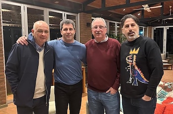 Dos hombres sentados frente a una mesa con dos vasos de agua, una servilleta, un posavasos tejido y un cuadro abstracto de tonos azules en la pared