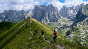 Un grupo de seis personas con mochilas y bastones camina por un sendero en la cresta de una montaña verde, con montañas rocosas al fondo y cielo con nubes.