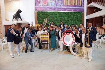 Grupo de veintisiete personas, incluyendo una mujer, posando con instrumentos musicales. Al fondo, una pared verde y un cartel de evento taurino. Escultura de toro negra