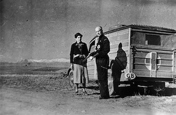 Fotografía en blanco y negro de Norman Bethune, un hombre calvo, y una mujer, de pie junto a una ambulancia antigua en un campo abierto con montañas nevadas al fondo