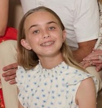 Una niña con ortodoncia y cabello claro sonríe con una blusa blanca de estampado floral azul