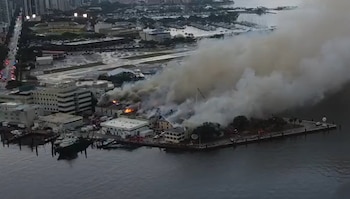 Vista aérea de un gran incendio en un complejo de edificios costeros con densas columnas de humo gris-marrón, vehículos de emergencia y un puerto deportivo
