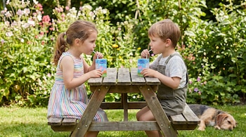 Un niño y una niña sentados en una mesa de picnic de madera en un jardín soleado, bebiendo de cartones de leche Peppa Pig. Un perro dormido en el césped.