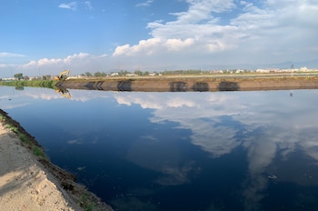 Vista panorámica de un canal de agua oscuro reflejando nubes, con un excavador amarillo en la orilla opuesta. En el fondo se ven árboles, una carretera y montañas