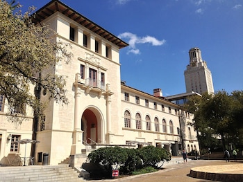 Edificio del Texas Union de piedra clara con entrada arqueada y ventanas, flanqueado por árboles y arbustos. La Torre de la UT se eleva al fondo bajo un cielo azul con nubes. Un sendero y personas caminando son visibles
