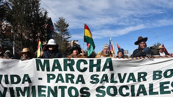 Un grupo de personas, algunas con sombreros, sostienen una pancarta blanca con texto verde. Banderas bolivianas y árboles son visibles bajo un cielo azul