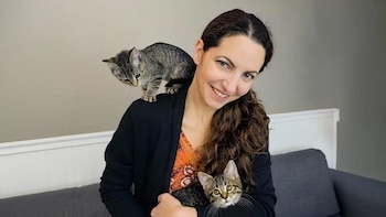Mujer de cabello largo y castaño con blusa naranja y cárdigan negro, sonríe a cámara. Sostiene un gatito atigrado y otro gatito atigrado está en su hombro