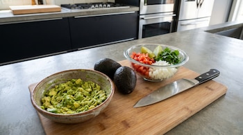 Aguacate triturado en tazón, dos aguacates enteros y otro tazón con tomate, cebolla, cilantro y limón en tabla de cortar con cuchillo en cocina moderna.