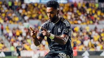 Un futbolista con barba y uniforme negro apunta con los dedos índices en un estadio con espectadores en el fondo.
