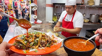 Un plato con tacos de carne, cebolla y cilantro, una cuchara vertiendo salsa, un tazón con salsa roja y un cocinero con delantal rojo.