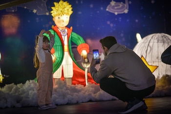 Una niña, un adulto fotografiando con un móvil, una figura iluminada del Principito, nubes de utilería y una pared azul con estrellas
