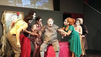 Seis actores maquillados y vestidos con trajes teatrales sobre un escenario. Un actor central es rodeado por otros. Hay un cuadro y una proyección de fondo