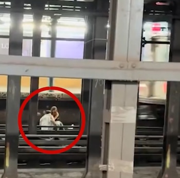 Dos personas sentadas sobre las vías en una estación subterránea, señaladas con un círculo rojo, ante columnas de acero y un tren en movimiento