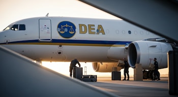 Avión blanco con logo de DEA y franjas azules y doradas en pista, tres agentes descargan cajas y maletas, amanecer cálido al fondo.