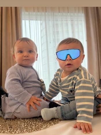 Dos bebés sentados uno al lado del otro frente a una ventana. El de la izquierda viste un mameluco lila. El de la derecha usa gafas de sol azules