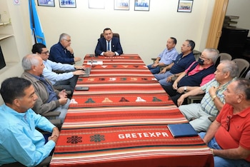 Nueve hombres sentados alrededor de una mesa rectangular con mantel rojo. Hay un portátil, teléfonos, un cuaderno y cuadros en la pared, junto a una bandera