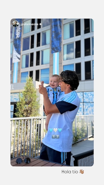 Hombre joven sostiene a un bebé sonriente, ambos con camisetas de Argentina. El bebé levanta un pulgar. Fondo: edificio con decoraciones celestes y blancas
