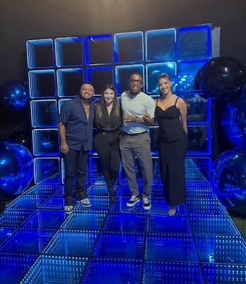 Cuatro personas posan de pie sobre una plataforma con luces azules frente a una pared de paneles y esferas oscuras