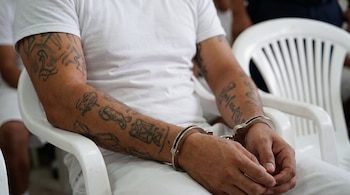 Hombre sentado en silla de plástico blanca, vestido de blanco, brazos tatuados, manos esposadas de metal.