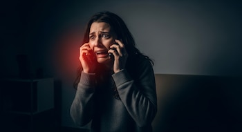Mujer de cabello oscuro y suéter gris, con expresión de pánico, sostiene un teléfono móvil en cada oreja. Fondo oscuro con una luz roja tenue a la izquierda.