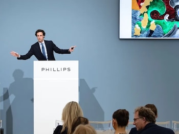 Hombre en traje gesticulando detrás de podio blanco con logo Phillips. Dos pantallas muestran obras de arte abstractas. Mujer y público sentados