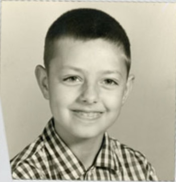 Fotografía en blanco y negro de un niño sonriendo, con el cabello corto y una camisa a cuadros. Fondo liso