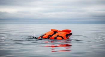 Un chaleco salvavidas naranja brillante flota en la superficie de un cuerpo de agua gris-azul con ligeras ondas, bajo un cielo nublado y sombrío.