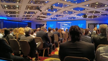 Vista trasera de una audiencia en un salón de conferencias con un orador en el escenario, frente a una pantalla azul brillante que muestra el logo "M'" y texto