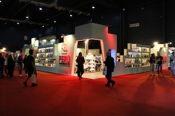 Pabellón interior con alfombra roja. Stand blanco iluminado con letras "PERÚ", exhibiendo libros. Varias personas caminan y observan los productos
