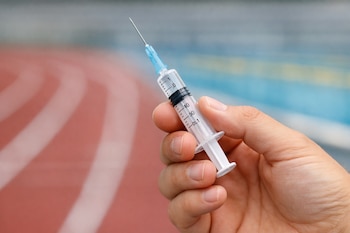 Plano detalle de una mano sosteniendo una jeringa con aguja y escala de medición, sobre un fondo desenfocado de una pista de atletismo o piscina.