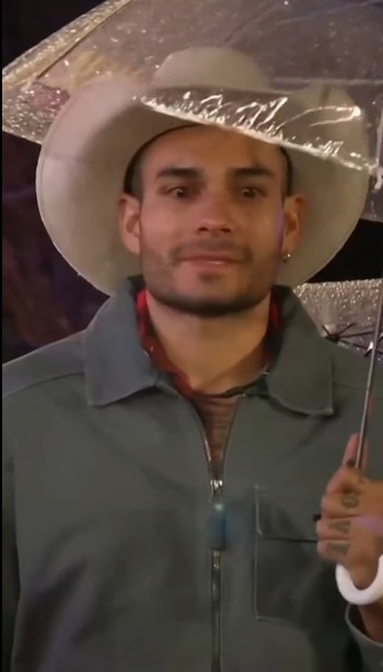 Un hombre con sombrero claro y chaqueta gris sostiene un paraguas transparente
