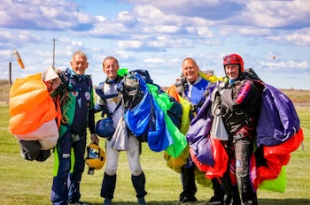 Cinco hombres con trajes de paracaidismo y paracaídas plegados de colores brillantes posan en un campo verde bajo un cielo azul nublado
