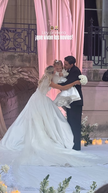 Cande Ruggeri, vestida de novia, y Nico Maccari, de traje, abrazan a su hija Ley, quien lleva un vestido blanco, frente a cortinas rosas en un evento al aire libre
