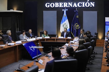 Grupo de hombres y mujeres sentados en una mesa de conferencia con micrófonos, botellas de agua y documentos. Detrás, dos banderas y un letrero que dice "GOBERNACIÓN"